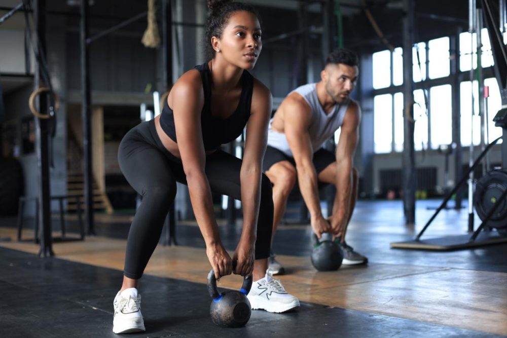 Deux femmes souriantes et sportives réalisant un biceps curl dans une salle de sport colorée.